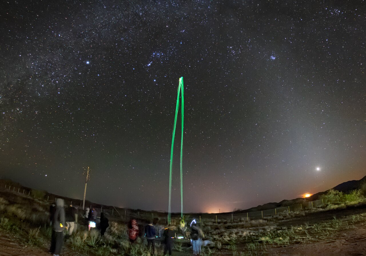 El personal de NOIRLab de NSF colabora a menudo con la sección de American Indian Science and Engineering Students (AISES) de la Universidad Comunitaria Tohono O'odham para organizar eventos de observación de estrellas y otras actividades locales. Una de las actividades más populares es que los estudiantes construyan y lancen cohetes de agua con botellas de 2 litros y barras luminosas para que los vuelos sean visibles en el oscuro cielo de Arizona. Esta imagen muestra la trayectoria de un lanzamiento que alcanza su ápice cerca de la constelación de Orión.